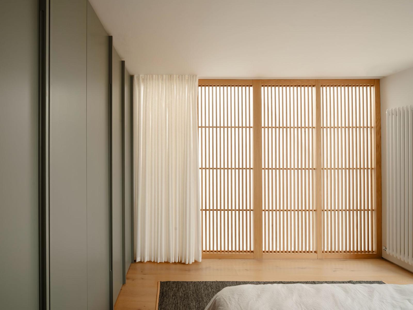 Contemporary bedroom with a slatted oak screen, sheer linen curtain and bespoke sage-green wardrobes