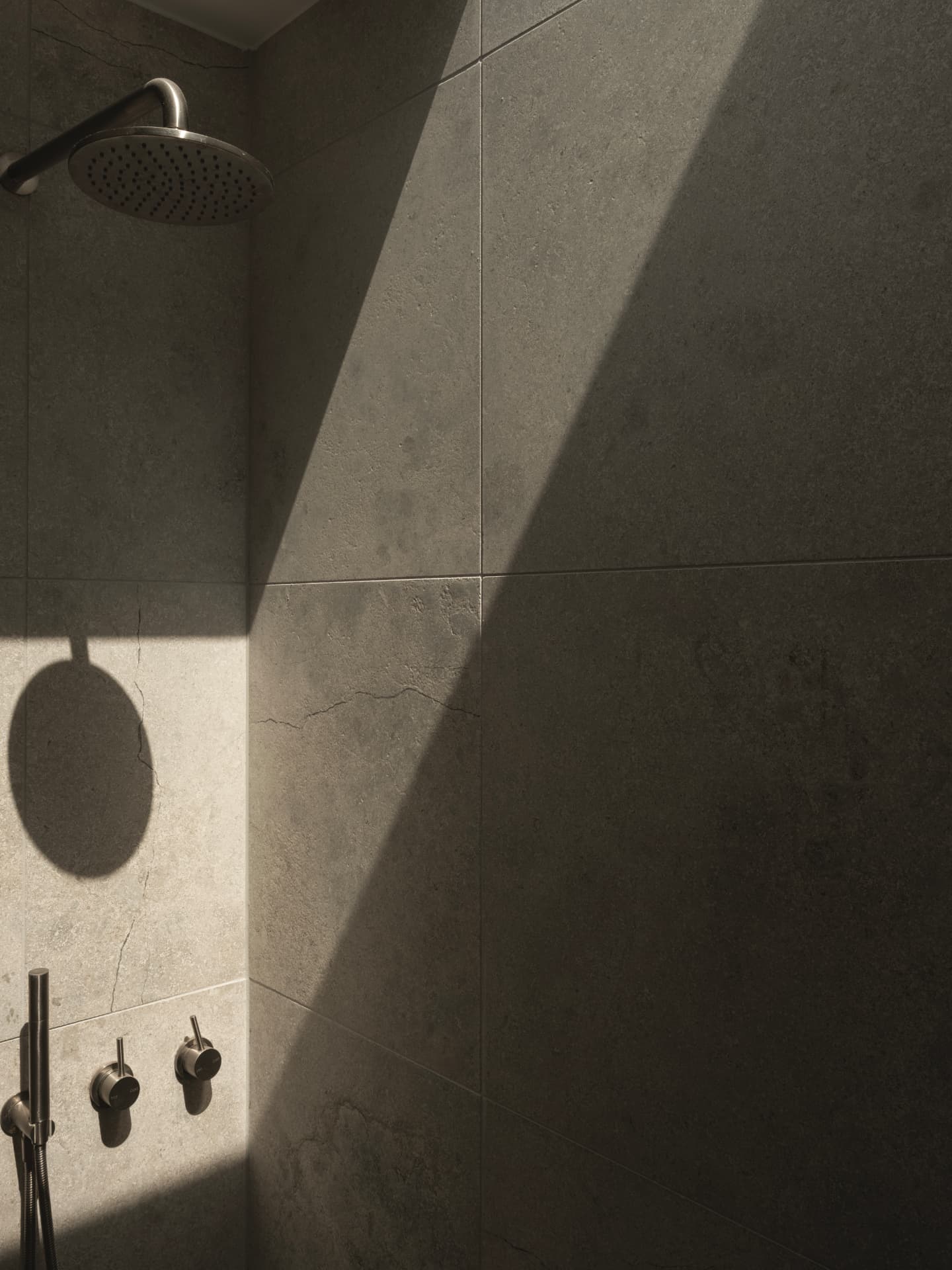 Brushed steel shower head casting a soft circular shadow across sunlit natural stone tiles
